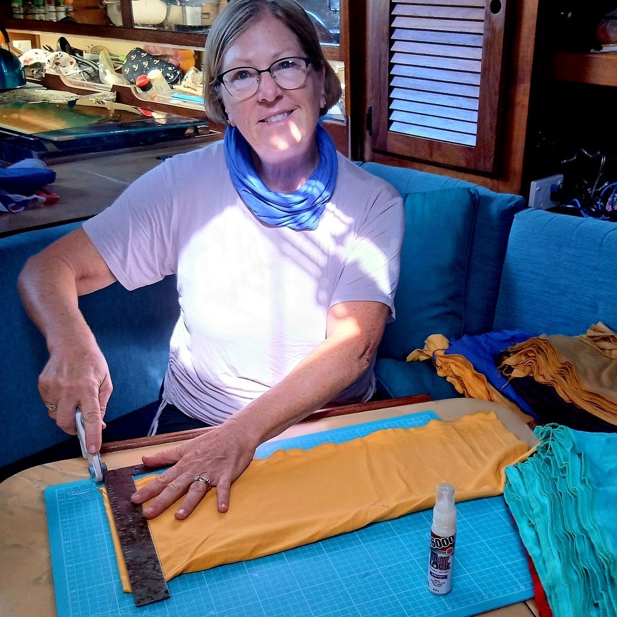 Charlotte on board, wearing a blue neck sock around her neck, cutting a length of orange fabric on a mat on the saloon table