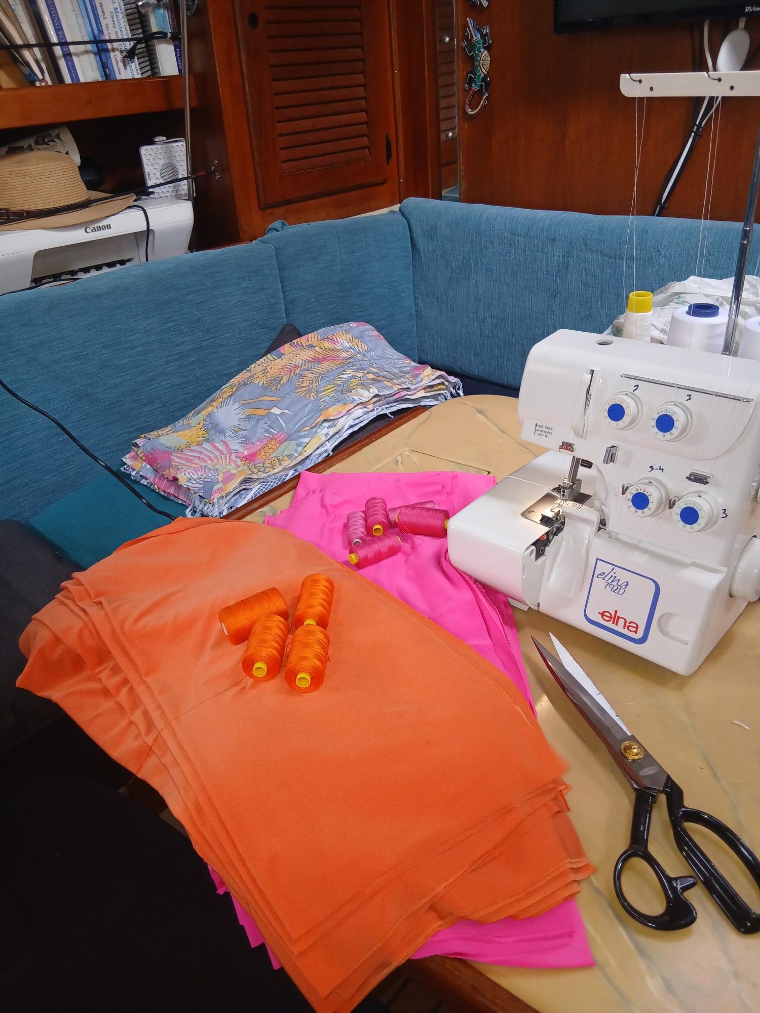 The saloon table set up for sewing: the overlocker, stacks of orange and pink fabric, spools of thread and a pair of shears