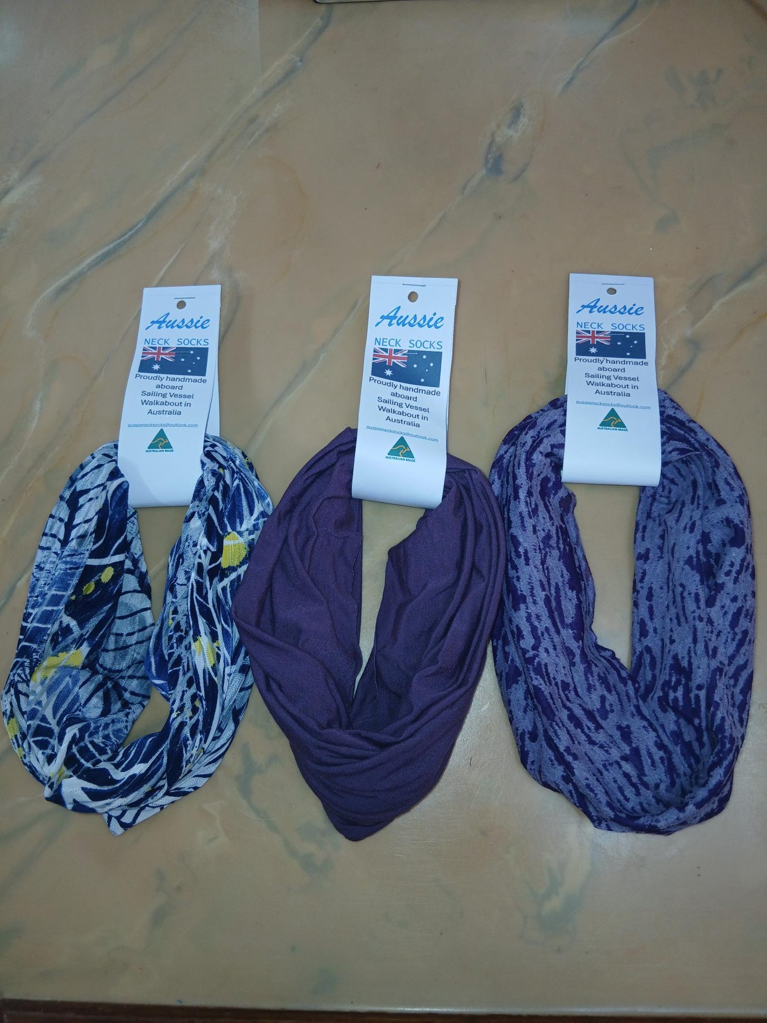 Three neck socks on the saloon table, each with its Aussie Neck Socks hangtag: a navy and yellow print, a plain plum and a purple pattern
