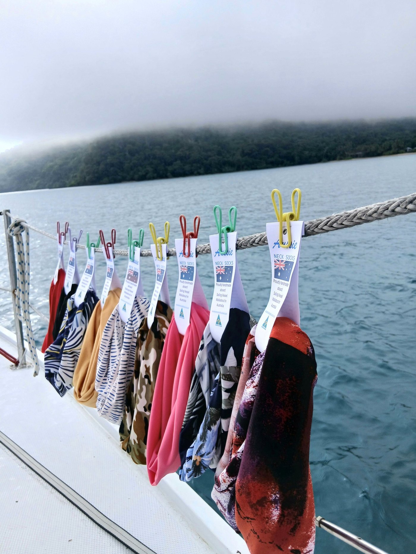 Neck socks in a row of bright colours, pegged along the lifelines of a sailboat to dry, with the water and a green headland behind